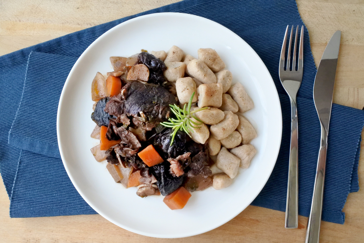 A plate of Pašticada featuring soft gnocchi and a stew, garnished with fresh rosemary, placed on a white dish over a blue napkin, with a knife and fork on the side.