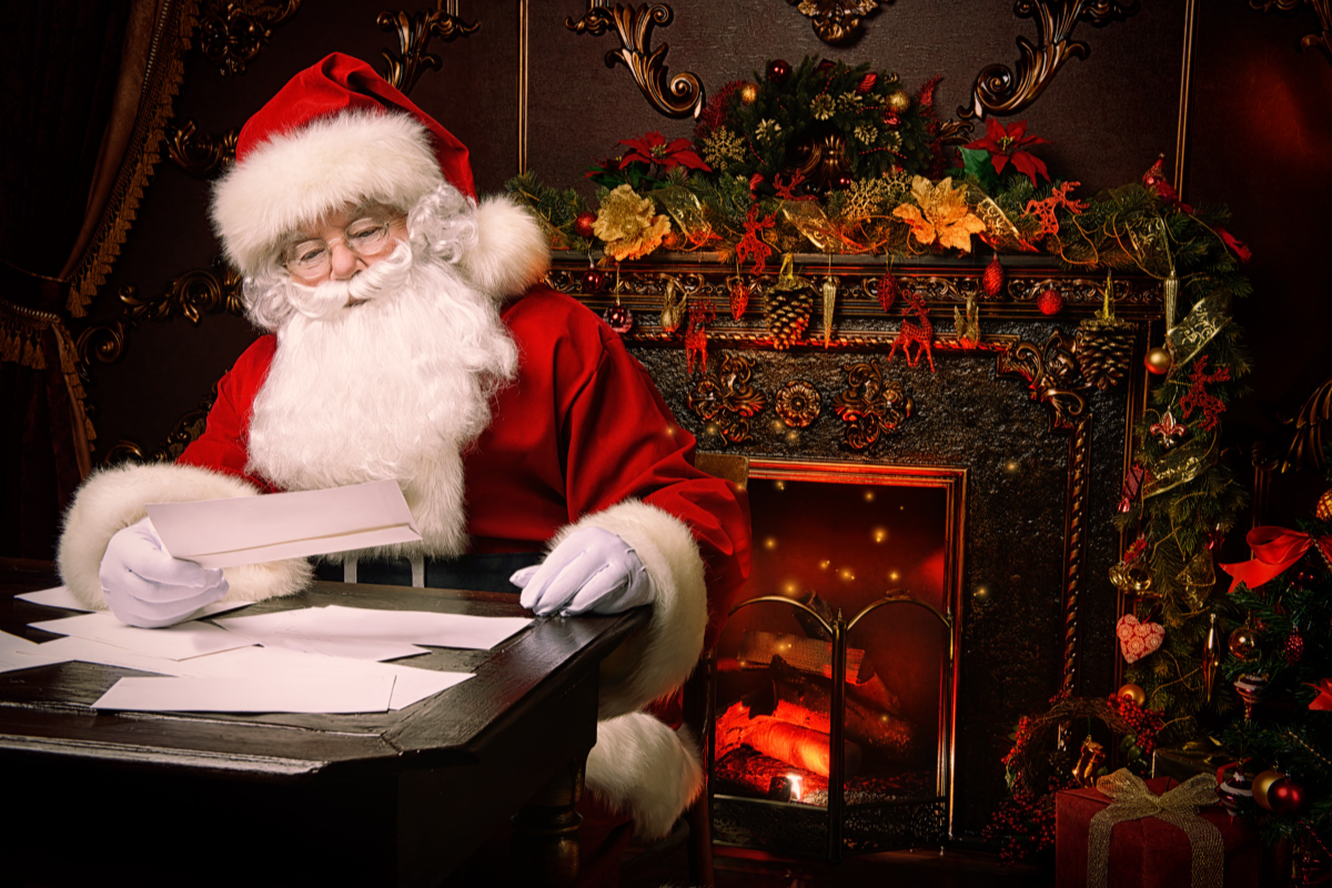 Santa reads children's letters in his living room with a lit fireplace