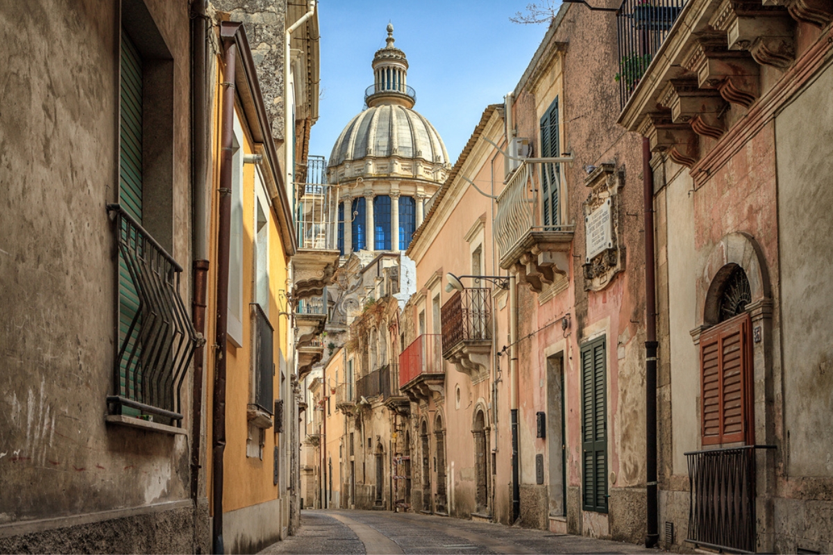 Narrow panoramic street in Ragusa, Sicily, Italy with old terraced houses and a church dome visible at the end, UNESCO World Heritage Site