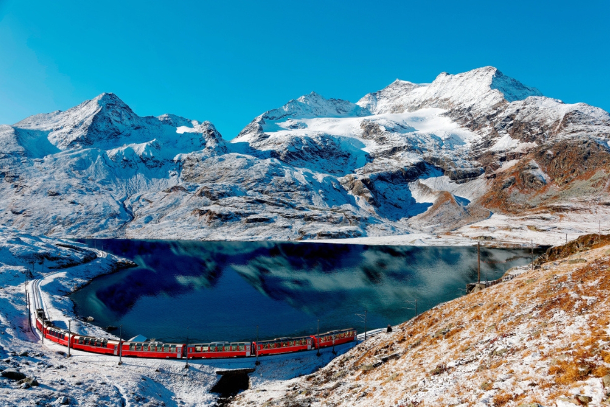 A Bernina Express train travelling along the shore of Lake Bianco and Piz Cambrena towering under the blue sky in the background after a snowfall, near Ospizio Bernina, Graubünden, Switzerland