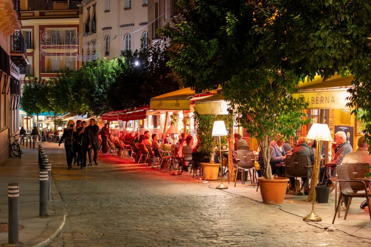 Una coloratissima strada di ciottoli fiancheggiata da alberi e da caffè illuminati di sera nel quartiere storico di Barrio Santa Cruz a Siviglia, in Spagna
