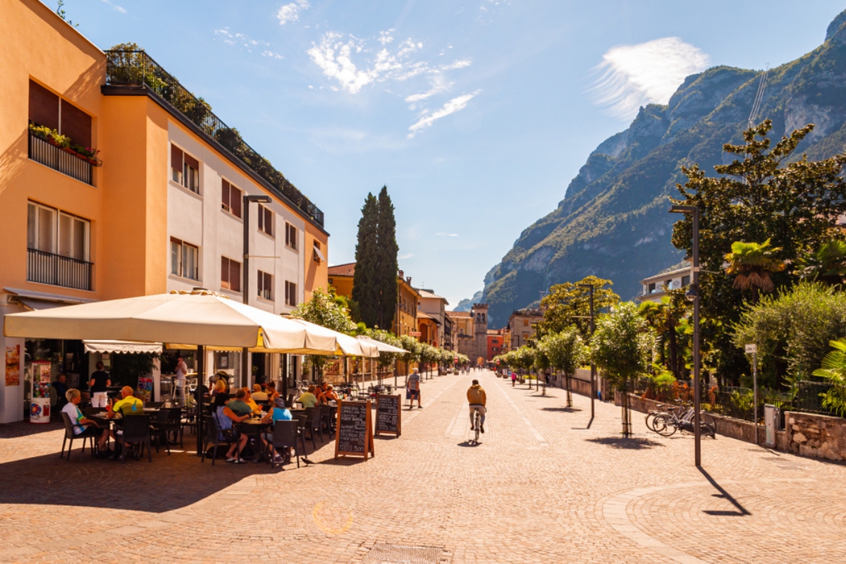 Scenic cityscape of Riva del Garda. Cosy city street full of tourists, plants and Italian architecture with high dolomite mountains on background