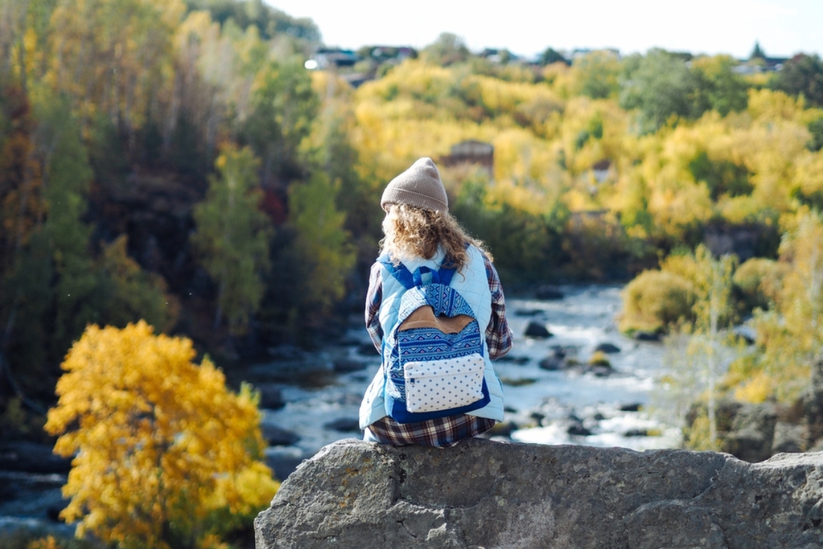 Young curly woman traveller in plaid shirt sitting on a rock; there is mountain river on a background