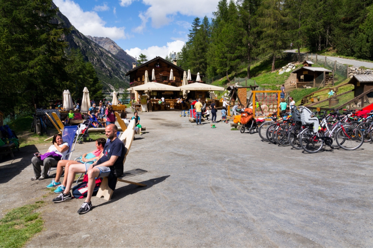 People sunbathing in front of restaurant with playing kids and bikes on bank of Lago di Livigno