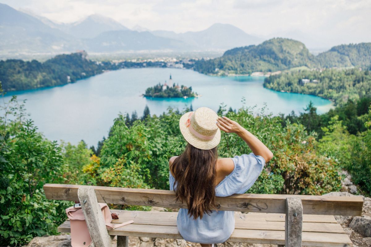 Donna in abito blu e cappello seduta su una panchina con davanti il lago di Bled in Slovenia