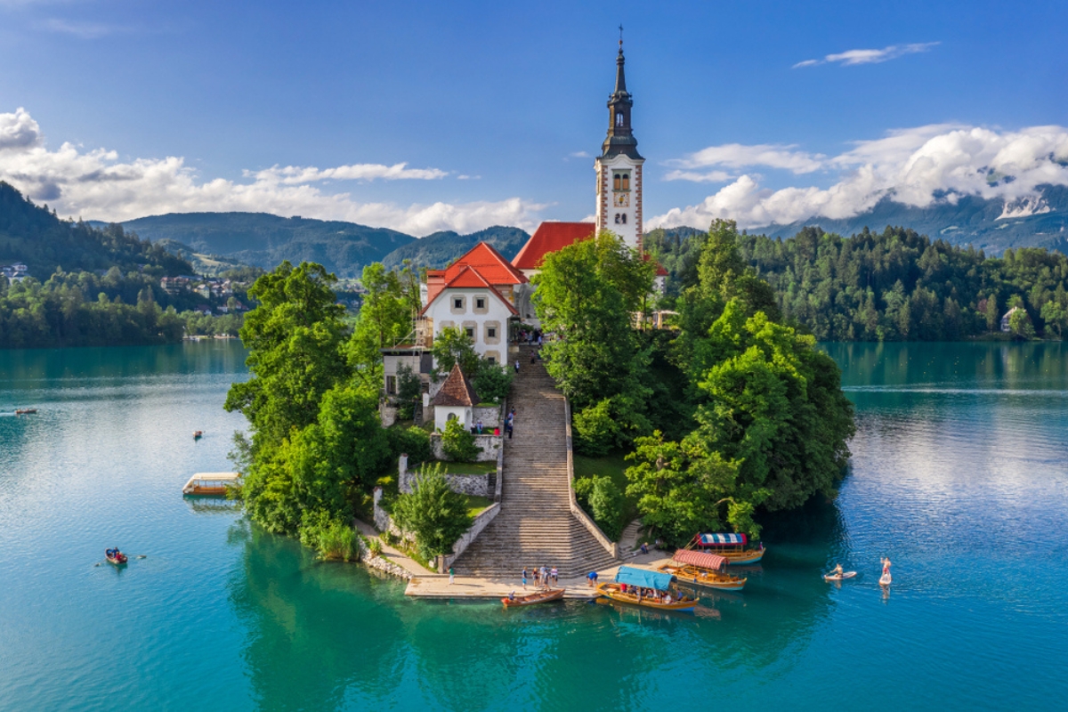 Bled, Slovenia - Veduta aerea della bellissima chiesa di pellegrinaggio dell'Assunzione di Maria su un'isoletta del lago di Bled (Blejsko Jezero) e di molte barche Pletna sul lago in estate con il cielo azzurro