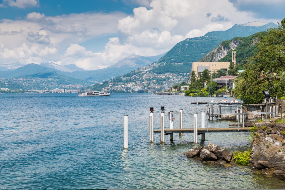 Lago di Lugano. Veduta di Campione d'Italia, famosa per il suo casinò (visibile sulla destra), con l'arrivo di un battello turistico. Sullo sfondo la città di Lugano e le Alpi svizzere.