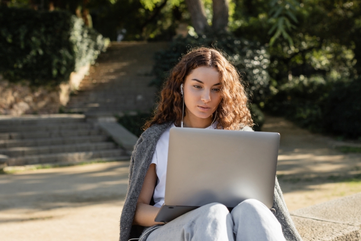 Giovane freelance con auricolari che utilizza il computer portatile mentre è seduta su un parapetto e trascorre il tempo in un parco a Barcellona, Spagna